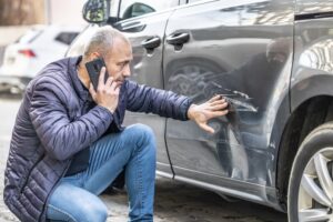 A man crouching by a damaged car after an accident, calling his insurance company to report the collision and assess damages exceeding insurance coverage.